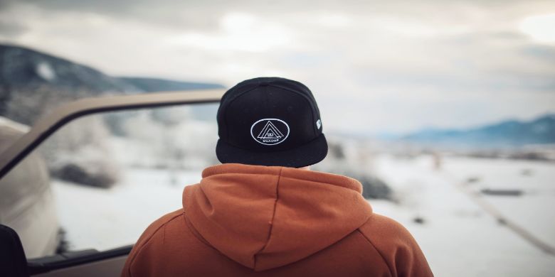 A guy wearing a cap in a brown jacket with snowy scenery.