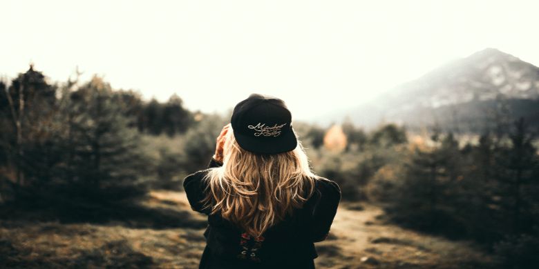 A blonde woman wearing their cap backwards in a forest-like location.